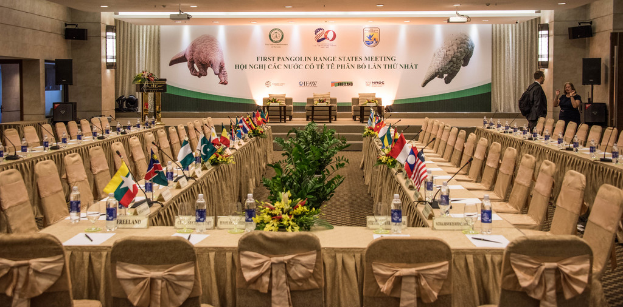 Eine B√ľhne mit Stühlen, Mikrofonen, Flaggen, Flaschen auf Tischen, einigen stehenden Menschen und einem Plakat.