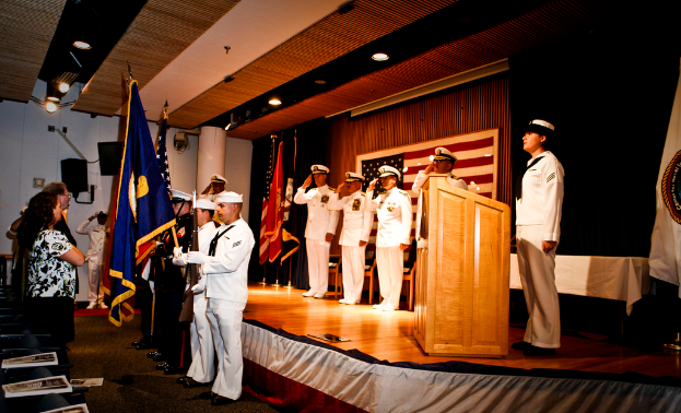 Eine formelle Veranstaltung mit Menschen, die auf einer Bühne stehen und salutieren, Flaggen in Rot, Blau und Weiß, Stühle, ein Podium und ein Tisch mit einer weißen Decke, mit einem Fernseher und einem Lautsprecherbox im Hintergrund.