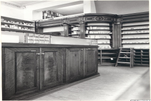 Ein Schwarz-Weiß-Foto einer Apotheken-Inneneinrichtung mit einem Tresen, Schränken, einer Leiter und Regalen, die mit verschiedenen Gegenständen gefüllt sind, mit Text oben drauf.