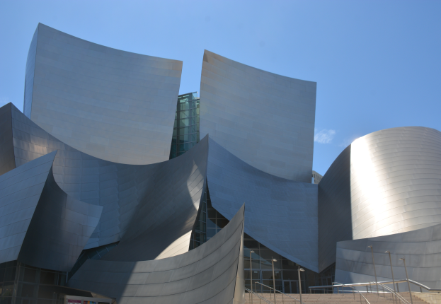 Das ARTIST-Konzertgebäude in Los Angeles, ein modernes Gebäude mit Glaswänden, Treppen mit Geländern, die zum Eingang führen, umgeben von Laternenmasten, mit einer Tafel mit Text im Vordergrund und einem bewölkten Himmel im Hintergrund.