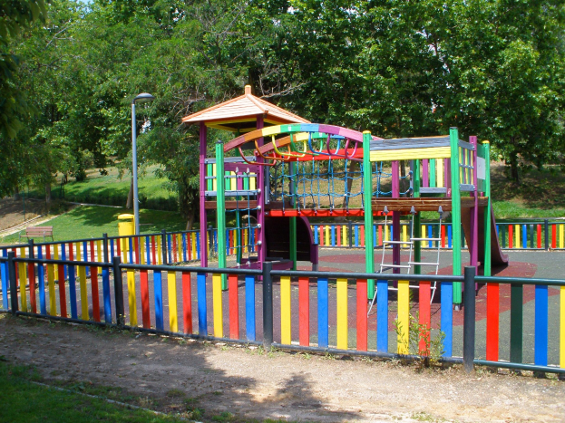 Ein Spielplatz mit einer bunten Spielstruktur in der Mitte, umgeben von einem Zaun, Pfosten, Laternen, Bänken, Gras, Pflanzen und Bäumen, unter einem klaren blauen Himmel.