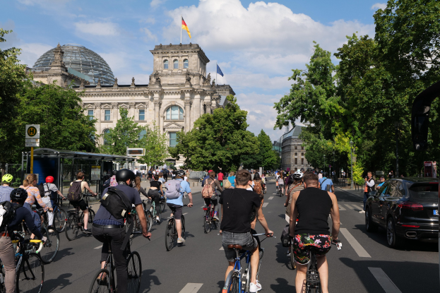 Eine Gruppe von Menschen, die auf Fahrrädern eine von Bäumen gesäumte Straße in Berlin, Deutschland, entlangfahren, mit Gebäuden und einer Bushaltestelle im Hintergrund, unter einem bewölkten Himmel und einer Flagge, die auf einem der Gebäude weht.