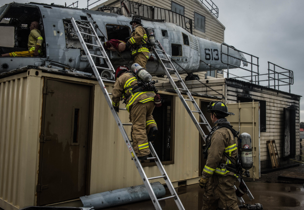 Feuerwehrleute in Uniformen und Helmen arbeiten an einem Hubschrauber auf einem Gebäudedach, mit Leitern, einem Sauerstoffzylinder, einem Container, Bäumen und einem klaren Himmel im Hintergrund.