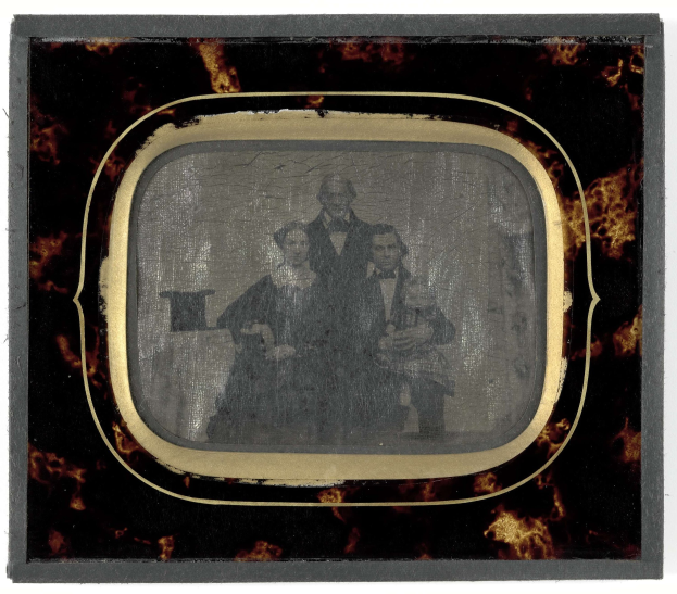 Altes Schwarz-Weiß-Foto eines Mannes im Anzug und einer Frau in einem unbestimmten Outfit, die nebeneinander vor einem Fernseher stehen und ernst schauen, vor einem weißen Hintergrund.