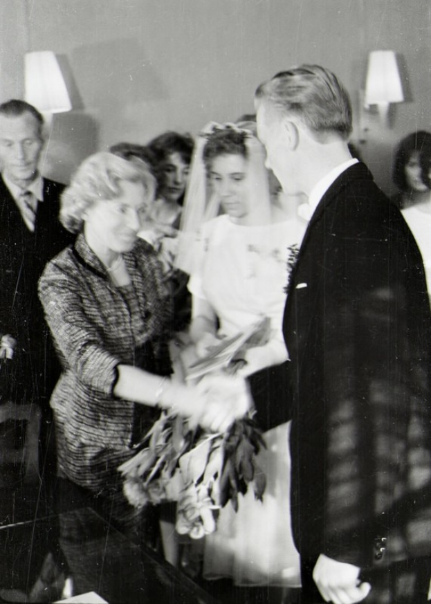 Ein Schwarz-Weiß-Foto eines Brautpaares beim Austausch der Ringe, wobei die Braut in einem weißen Kleid und Schleier mit einem Strauß Blumen in der Hand zu sehen ist und der Bräutigam in einem Anzug und Krawatte, mit Wandlampen im Hintergrund.