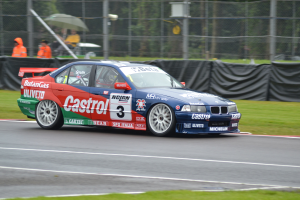 Ein Auto auf einer Rennstrecke mit Text und Zahlen, umgeben von Gras, mit Maschendrahtzaun, Menschen in orangefarbenen Jacken mit Schirmen und Bäumen im Hintergrund.