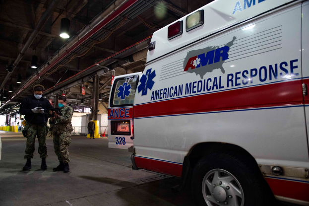 Ein Rettungswagen der American Medical Response (AMR) steht in einem Gebäude mit zwei maskierten Personen davor, einem Fahrzeug auf der linken Seite und mehreren Personen, Lampen und Rohren im Hintergrund.
