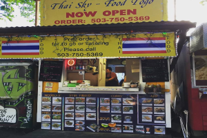 Ein Schaufenster mit verschiedenen Artikeln wie Getränkedosen, Speisekarten, einer Uhr, Gewürzen in Plastikbehältern, Gewürzen, Werbetafeln und Namensschildern unter einem Himmel.