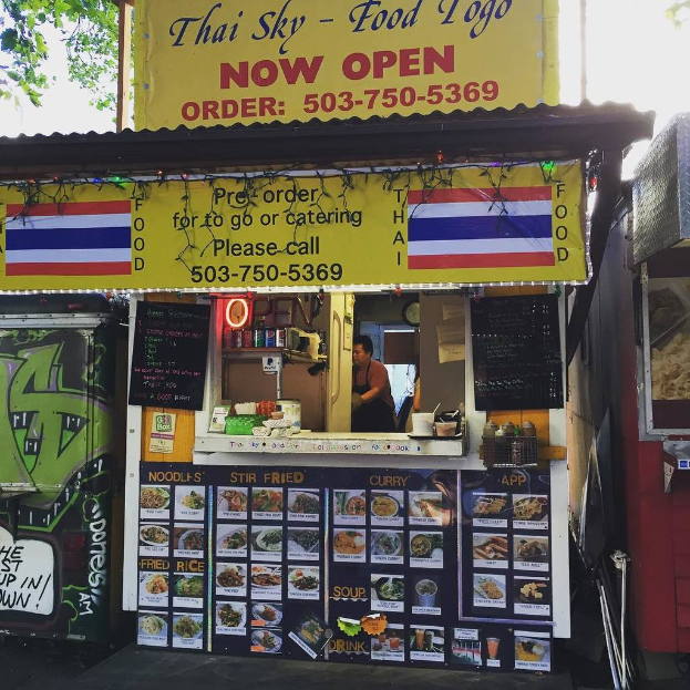 Ein Schaufenster mit verschiedenen Artikeln wie Getränkedosen, Speisekarten, einer Uhr, Gewürzen in Plastikbehältern, Gewürzen, Werbetafeln und Namensschildern unter einem Himmel.