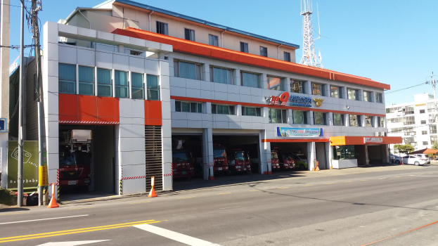 Feuerwache mit einem Feuerwehrauto davor geparkt, umgeben von Gebäuden, Fahrzeugen, Verkehrskegeln, Strommasten, Kabeln, Schildern, Bäumen und einem klaren Himmel.