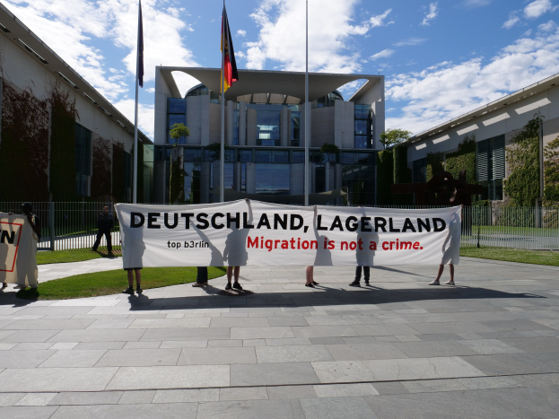 Gruppe von Menschen mit einer Fahne mit der Aufschrift "Deutschland, Lagerland Migration ist kein Verbrechen" vor einem Gebäude mit Fenstern, umgeben von Gras, einem Metallzaun, Pflanzen, Bäumen und Fahnen auf Stangen unter einem bewölkten Himmel.