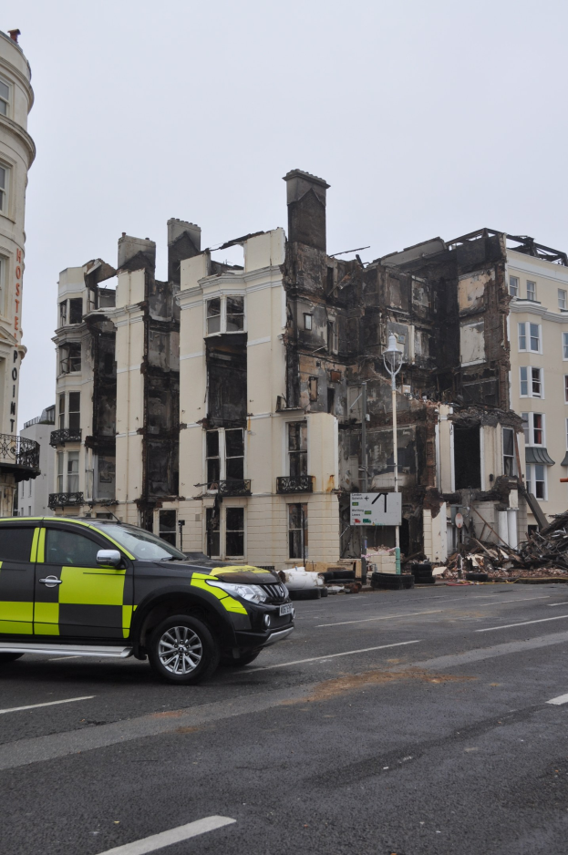 Ein Polizeiauto vor einem stark beschädigten Gebäude mit zerschlagenen Fenstern und verstreuter Trümmer, unter einem sichtbaren Himmel mit nahen Gebäuden.