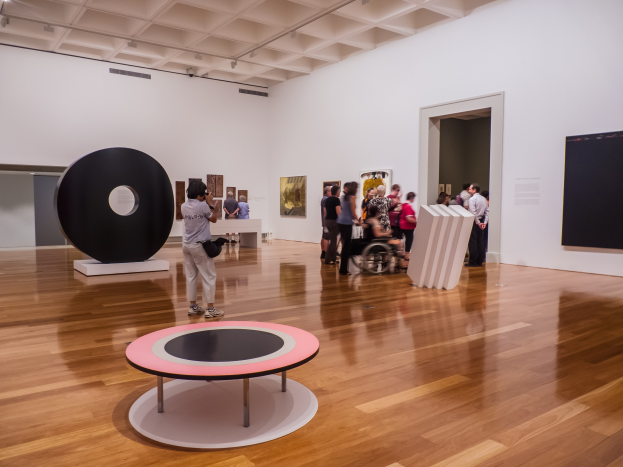 Eine Gruppe von Menschen in einer Kunstgalerie mit einer Person im Rollstuhl in der Mitte, umgeben von gerahmten Fotos an den Wänden und Deckenleuchten, neben einem schwarzen Installationsobjekt, das als Teil der Ausstellung 'Emanationen: Die Kunst der kameralosen Fotografie' an der Govett-Brewster Art Gallery gekennzeichnet ist.