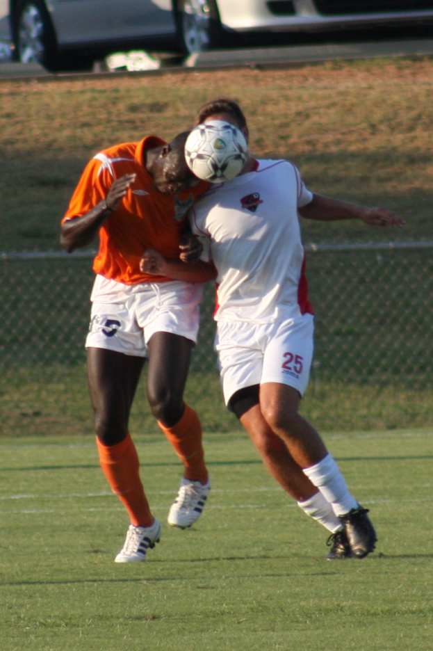 Zwei Spieler auf einem umzäunten Platz, einer im orangen T-Shirt schießt den Ball mit dem Kopf, mit Autos und Gras im Hintergrund.