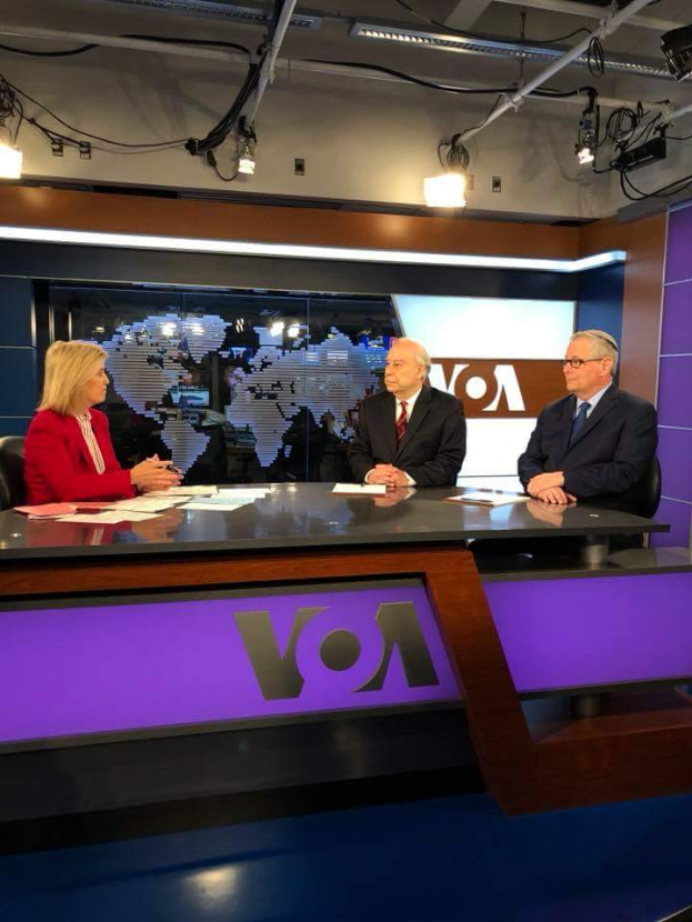 Drei Personen, zwei Männer in Blazern und Krawatten und eine Frau in einem roten Kleid, sitzen an einem Schreibtisch mit Papieren und Gegenständen, diskutieren vor einem Fernseher mit einem Bildschirm und Lampen im Hintergrund.