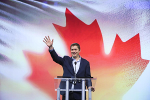 Kanadischer Premierminister Alex Salmond steht hinter einem Rednerpult mit Mikrofonen, lächend vor einer kanadischen Flagge, mit einem Bildschirm im Hintergrund.