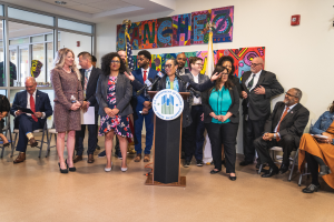 Eine Gruppe von Menschen, die vor einem Podium mit Mikrofon und einer Tafel mit der Aufschrift "Sancho City Councilman Joins City Council" stehen und sitzen, mit Flaggen, Feuerl├Âscher und Deckenleuchten an der R├╝ckwand.