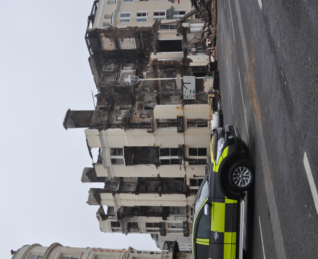 Ein Polizeiwagen steht vor einem zerstörten Gebäude mit zerschmetterten Fenstern und verstreuter Trümmer, mit anderen Gebäuden und dem Himmel im Hintergrund.