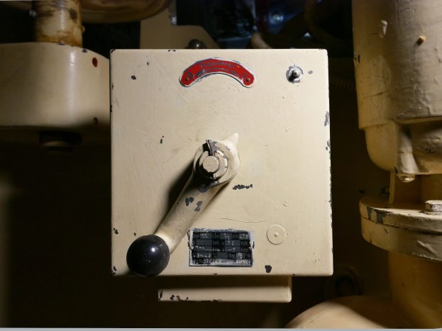 Nahaufnahme einer Steuerkonsole mit Drehknopf und Messgerät, umgeben von Rohren.