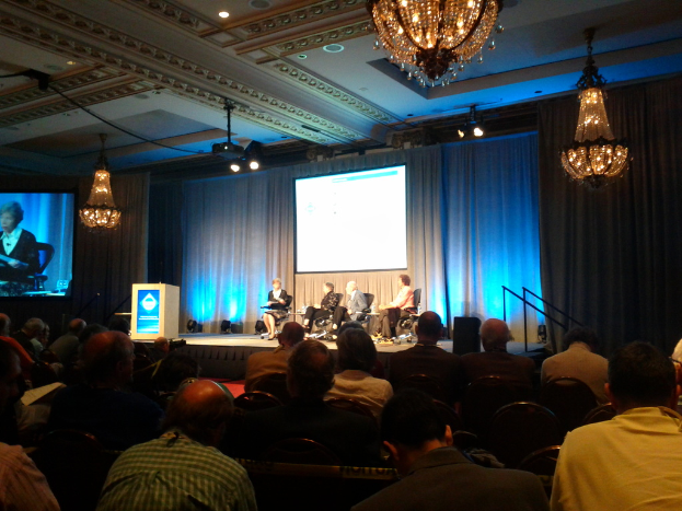 Eine Gruppe von Menschen, die auf Stühlen auf einer B"{u}hne mit einem Podium, Lichtern, Bildschirmen, Vorh"{a}ngen, Kronleuchtern, einem Lautsprecher und einem Dach sitzen.