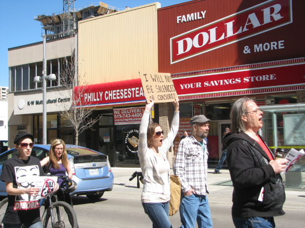 Eine Gruppe von Menschen protestiert auf einer Straße mit Gebäuden, einem Pfahl und einem Baum im Hintergrund, einem Auto und einem Fahrrad auf der linken Seite und dem Himmel oben.