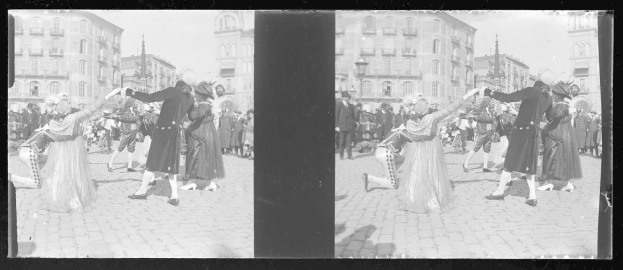 Ein Schwarz-Weiß-Foto einer Gruppe von Menschen, die auf der Straße tanzen, während eines Karnevalsumzugs, gekleidet in Kostüme, mit Gebäuden, Straßenlaternen und Straßenlaternen im Hintergrund unter einem klaren Himmel.
