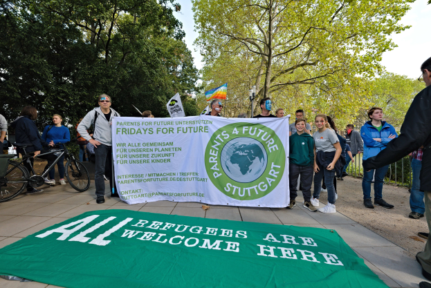 Gruppe von Menschen mit einer Fahne und einem Banner mit der Aufschrift "All Refugees Are Welcome Here", umgeben von Fahrrädern, einem Zaun, einem Straßenschild, einem Schild, Bäumen und einem bewölkten Himmel.
