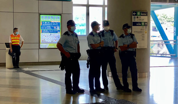 Eine Gruppe maskierter Polizisten steht vor einem Gebäude mit Glastüren, Texttafeln, einer Säule und einem Geländer, wobei durch die Türen Bäume und Fahrzeuge zu sehen sind.