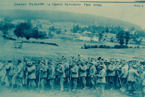 Eine Gruppe deutscher Gefangener in Frankreich, die Waffen haltend auf dem Boden stehen, mit Bäumen, Häusern und einem klaren blauen Himmel im Hintergrund.