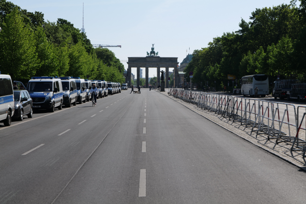 Eine Reihe von Polizeiwagen, die auf der Seite einer Straße vor dem Brandenburger Tor in Berlin, Deutschland, geparkt sind, mit Menschen, die Fahrräder fahren und auf der Straße stehen, sowie Barrieren und Bäume, die die Seiten säumen, und einem Bogen mit Statuen im Hintergrund.