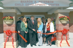 Eine Gruppe von Menschen steht vor dem Sharon A Peterson Child Development Center, einige tragen Kränze und einer hält eine Tasche, mit Bändern an den Säulen und Blumentöpfen auf beiden Seiten.
