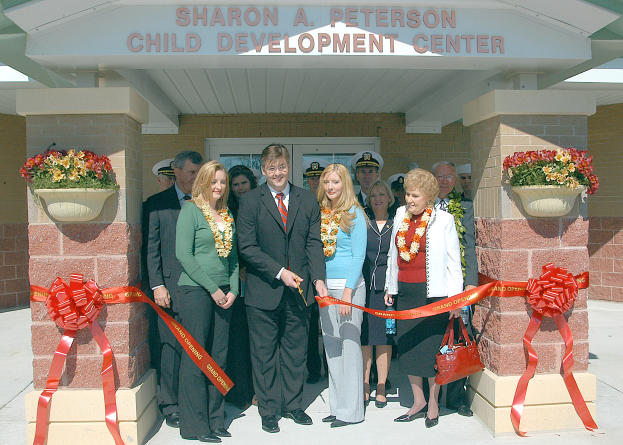 Eine Gruppe von Menschen steht vor dem Sharon A Peterson Child Development Center, einige tragen Kränze und einer hält eine Tasche, mit Bändern an den Säulen und Blumentöpfen auf beiden Seiten.