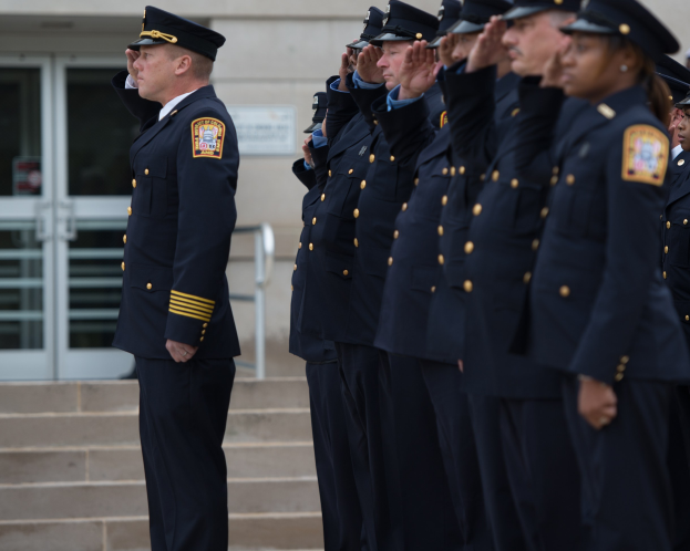 Gruppe von Polizisten in Uniform und Mützen in Formation vor einem Gebäude mit Glastüren und Treppe stehend und salutierend.