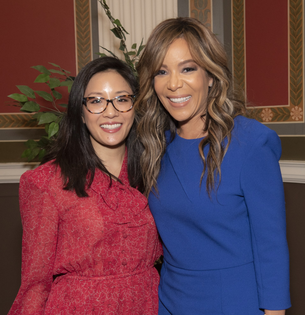 Zwei Frauen stehen nebeneinander und lächeln, eine trägt ein blaues Kleid und die andere ein rotes Kleid mit Brille, auf der 2019 Women in Film Pre-Oscar-Luncheon mit einer Pflanze und einem Vorhang im Hintergrund.