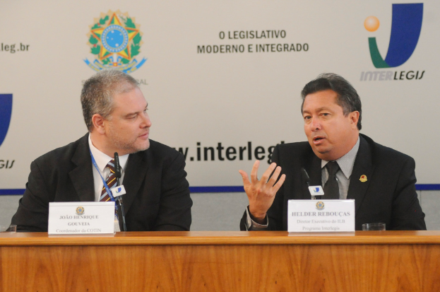Zwei Männer in Anzügen sitzen an einem Tisch mit zwei Namensschildern und zwei Mikrofonen, mit Text und Logos im Hintergrund.