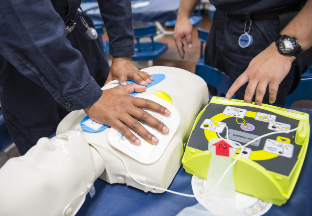 Zwei Polizeibeamte neben einer Puppe auf einem Tisch in einer hospitalähnlichen Umgebung, mit Defibrillator-Trainingsequipment sichtbar.