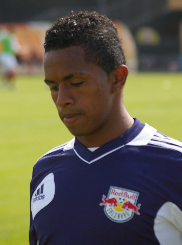 Ein junger Mann in einem blauen T-Shirt steht auf einem Fussballfeld, mit kurzen dunklen Haaren und einem entschlossenen Gesichtsausdruck, der als Flügelspieler mit einem unscharfen Hintergrund erscheint.