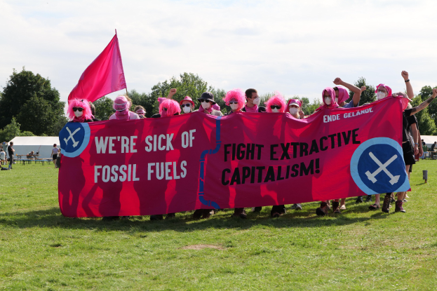 Eine Gruppe von Menschen steht auf einer grünen Wiese und hält ein pinkes Banner mit der Aufschrift "Wir haben genug von fossilen Brennstoffen, kämpfen gegen den extraktiven Kapitalismus", mit Zelten, Bäumen und einem klaren blauen Himmel im Hintergrund.