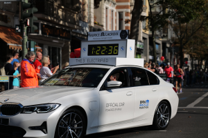 Ein weißer BMW mit einer Uhr auf dem Dach fährt eine Straße entlang, mit einer Person darin und einer Gruppe von Menschen, die auf der Straße stehen; Gebäude, Bäume und Ampeln sind im Hintergrund zu sehen.