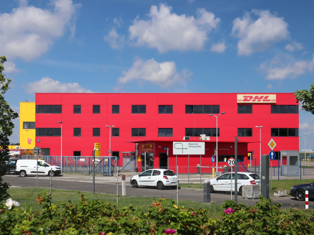 Ein rotes DHL-Gebäude in Deutschland mit parkenden Autos, umgeben von Pflanzen, Gras, einem Zaun, Schildern, Straßenlaternen, Bäumen und einem bewölkten Himmel.