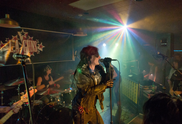 Eine Frau in einer schwarzen Jacke singt in ein Mikrofon auf der Bühne, während ein Mann mit Sonnenbrille und Stirnbinde ein Instrument spielt; ein "LA Guns"-Plakat und Diskoleuchten sind im Hintergrund zu sehen.