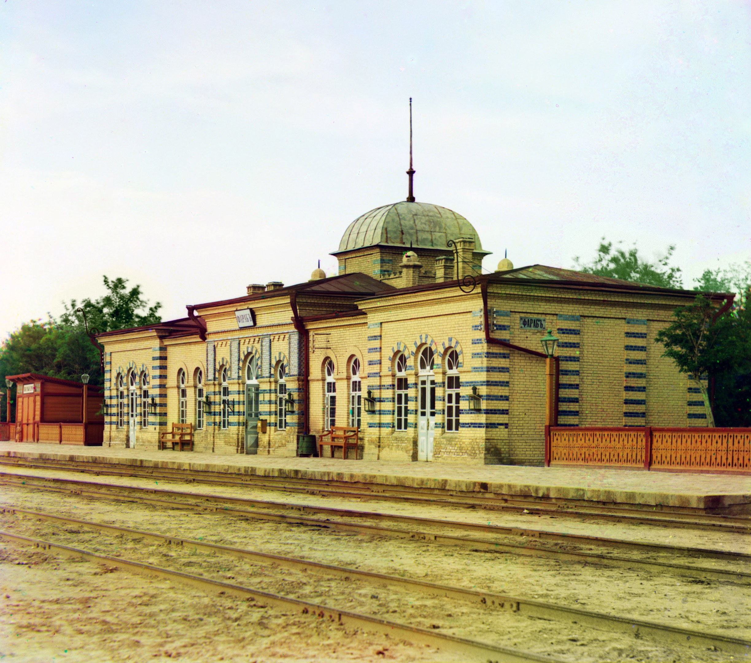 Altes Schwarz-Weiß-Foto eines Bahnhofs mit einem Gebäude, Bänken, einem Zaun, Laternen, Bäumen, einem bewölkten Himmel und einer Bahnstrecke unten.
