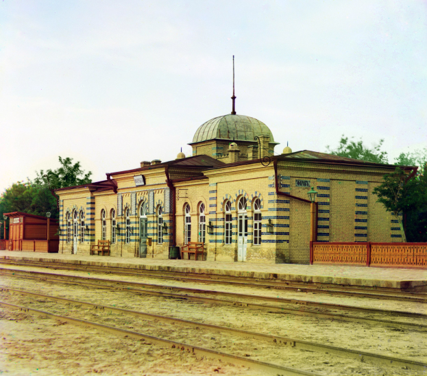 Altes Schwarz-Weiß-Foto eines Bahnhofs mit einem Gebäude, Bänken, einem Zaun, Laternen, Bäumen, einem bewölkten Himmel und einer Bahnstrecke unten.