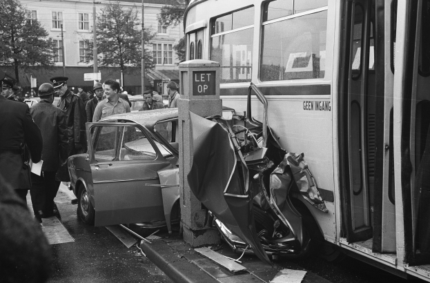 Schwarz-weißes Bild eines Busses, der auf der Straße in ein Auto gekracht ist, mit Menschen um die Unfallstelle und Bäumen und Gebäuden im Hintergrund.