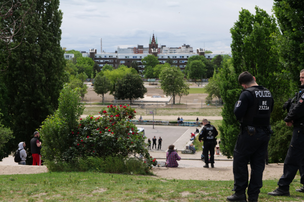 Zwei Polizeibeamte vor einer Gruppe von Menschen in einem Park mit Grünflächen, bunten Blumen, Gebäuden und einem klaren blauen Himmel.