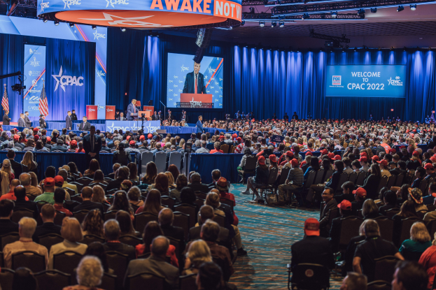 Eine große Menschenmenge sitzt auf Stühlen und schaut auf eine Bühne, auf der ein paar Menschen auf dem Podium und auf der linken Seite stehen, blaue Vorhänge und ein Bildschirm im Hintergrund und eine orangefarbene Tafel oben, die das CPAC 2020-Event am 25. Oktober anzeigt.
