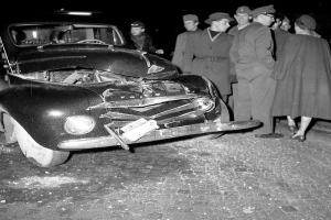 Ein Schwarz-Weiß-Bild eines auf der Straße liegenden, stark beschädigten Autos, um das sich eine Gruppe von Menschen schart.