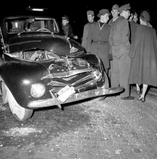 Ein Schwarz-Weiß-Bild eines auf der Straße liegenden, stark beschädigten Autos, um das sich eine Gruppe von Menschen schart.