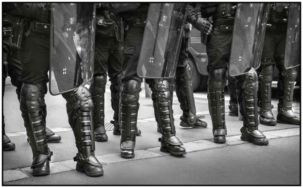 Schwarzes und weißes Bild einer Reihe von Polizisten mit Schilden, mit einem Fahrzeug im Hintergrund.