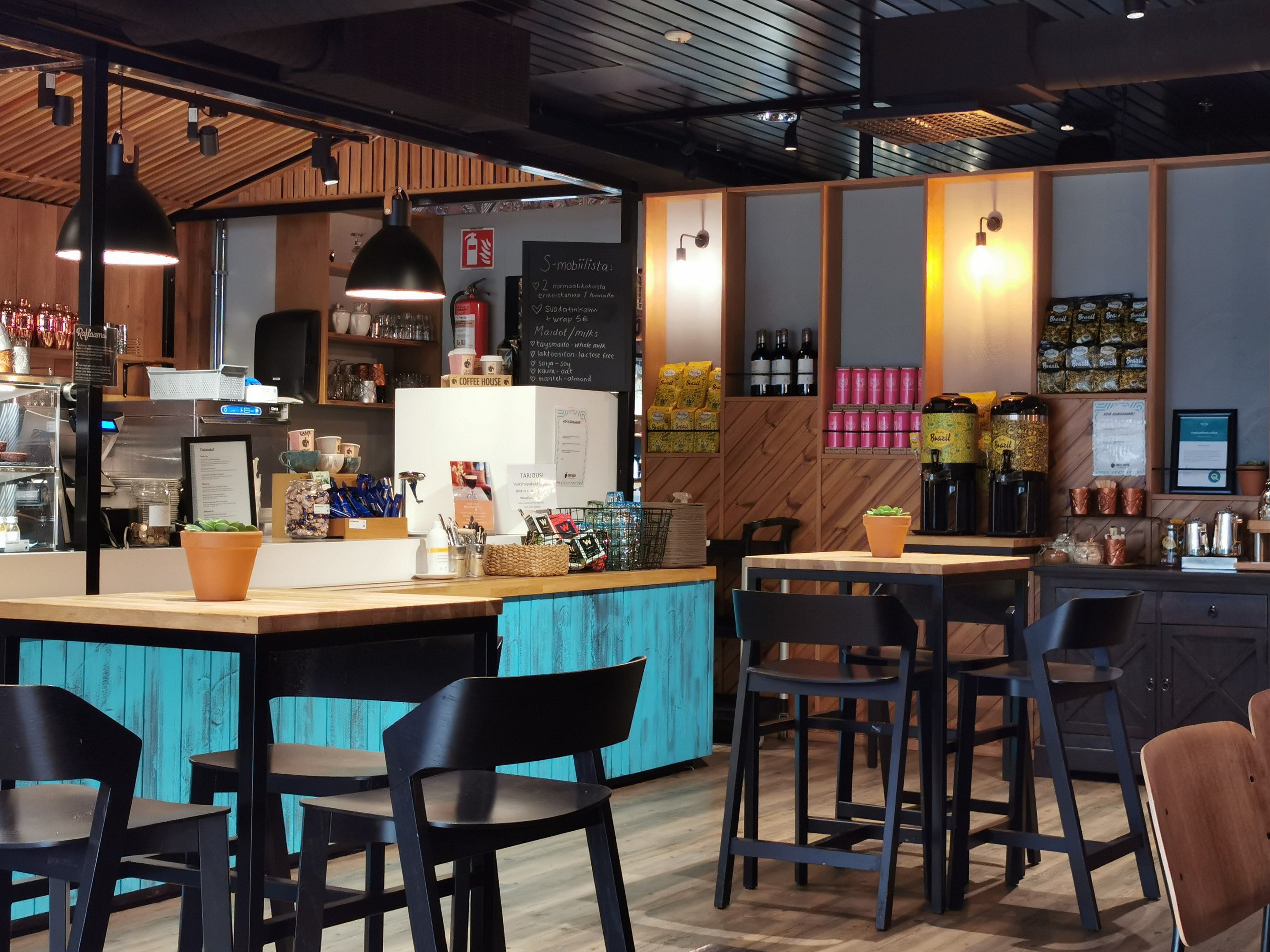 Innenraum eines Restaurants mit Holztischen, Stühlen, einer Topfpflanze auf einem Tisch, einer Schiefertafel, einem Kühlschrank, einem Feuerlöscher, Regalen mit Flaschen, Schränken mit Gegenständen, einem gerahmten Foto an der Wand und Deckenleuchten.